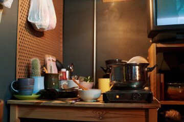 Corner of kitchen in small apartment with metallic pan standing on electric stove surrounded by bowls, mugs and other kitchenware