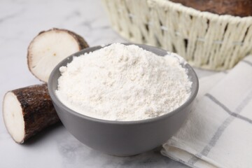 Bowl with cassava flour and roots on white marble table, closeup