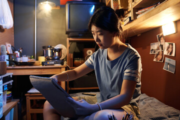 Chinese girl sitting on bed in front of camera in small apartment and making sketch in notepad...