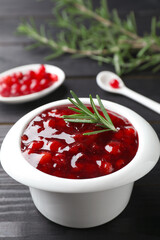 Fresh cranberry sauce in bowl and rosemary on black wooden table, closeup