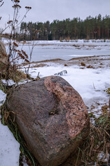 Winter landscape with big granite boulder on foreground and snow covered lake near coniferous forest
