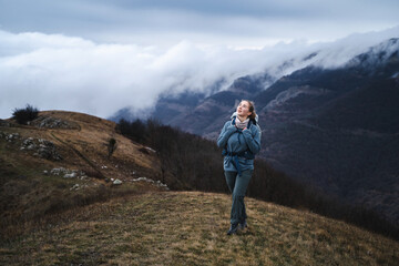 A happy young girl walks and laughs in hiking clothes on the top of a mountain above the clouds, a hiker on the way is filled with emotions and enjoys the beauty of the landscapes, in full view