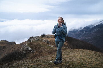 A young girl mountaineer walks and laughs in hiking clothes on the top of a mountain above the clouds, hiker having trekking day out on a windy day, sport and inspirational concept