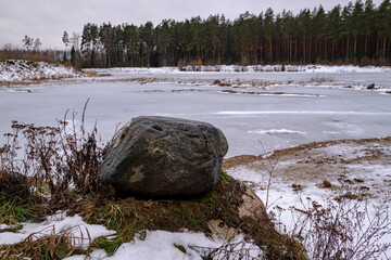 Winter landscape with big granite boulder on foreground and snow covered lake near coniferous forest