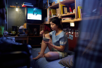 Young Asian woman with chopsticks in bowl of Chinese food sitting on bed in small apartment,...