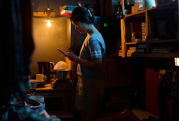 Low key image of young Asian woman with mobile phone standing by electric stove in the kitchen of microflat and preparing lunch