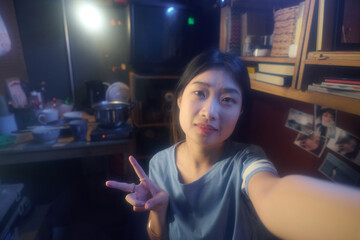 Happy young Chinese woman showing peace gesture and looking at camera while taking selfie or making livestream from microflat