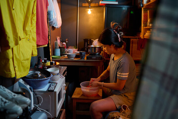 Side view of young woman sitting on bed against kitchen table in small apartment and washing towel or her clothes in plastic basin
