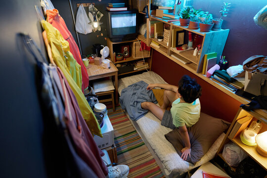 High angle of young Asian woman in casualwear sitting on single bed in front of screen of old TV set and watching broadcast or movie