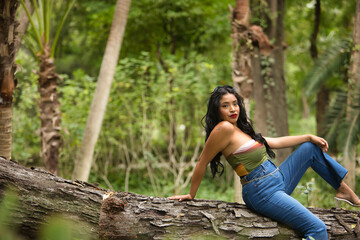 South American woman, young, beautiful, brunette, with colorful top and jeans sitting on a tree trunk, with sensual and provocative attitude. Concept of beauty, fashion, trend, ethnicity, diversity.