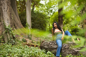 South American woman, young, beautiful, brunette, with colorful top and jeans sitting on a tree trunk, with sensual and provocative attitude. Concept of beauty, fashion, trend, ethnicity, diversity.
