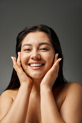 joyful and young asian woman with brunette hair touching acne prone skin on grey background