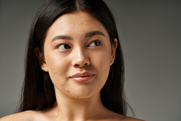 dreamy asian young woman with brunette hair and bare shoulders looking away on grey background
