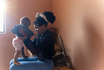 village african girl with braids, holding a baby she is sited next to the window indoors