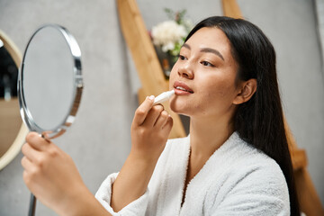 young and pretty asian woman in bath robe applying lip balm and looking at mirror in bathroom - obrazy, fototapety, plakaty