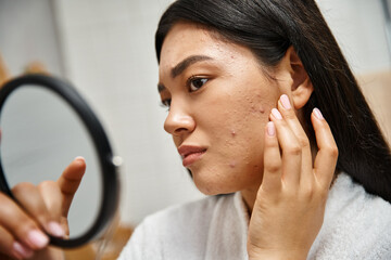 young asian woman with brunette hair and pimples looking at her face in mirror, skin issues