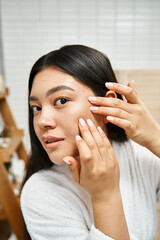 young and brunette asian girl in bath robe popping pimple on her face, skin issues or real people