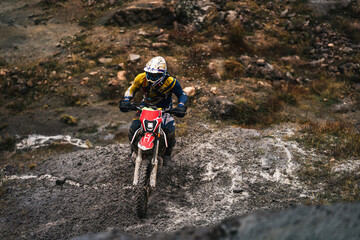 Portrait of professional motorcyclist in full moto equipment riding crops enduro bike in mountains,...
