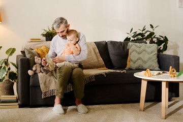 Mature man in casualwear sitting on couch in living room and holding cute baby son on knees while...