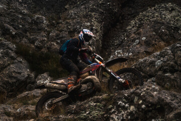 A racer at a competition rides along a cliff in rainy weather, bottom view, professional motorcyclist in full moto equipment riding crops enduro bike in highlands, concept of motosport