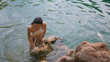 Swimming lady climbing rock deep lake. Young woman sitting brown volcanic stones