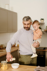 Mature father with his adorable baby son on hands standing by kitchen counter and pouring fresh milk into bowl with cornflakes