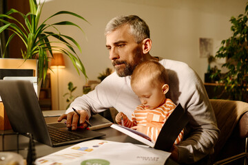 Businessman in casualwear looking at laptop screen while sitting by table and holding his cute baby...