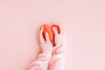 Hands hold red origami in the shape of a red heart on a pink background.