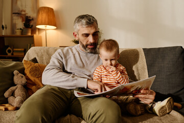 Mature father pointing at picture on page of open book of comics or fairy tales while reading it to...