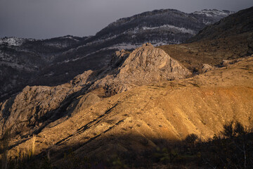 Landscape of rocks at classic golden hour glow, sunrise in snowy mountains, dramatic gray sky, snowy forest and yellow sun light, layers of mountains