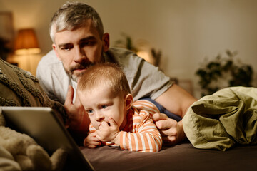 Obraz premium Mature father lying on bed next to sweet baby son while both looking at tablet screen and watching online cartoons in the evening