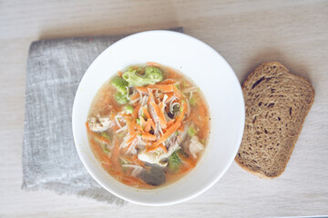 Top view of a healthy and delicious soup of vegetables and meat in a white plate on the table with bread with copy space. Bright soup with carrots broccoli, pieces of meat, served in plate with bread.