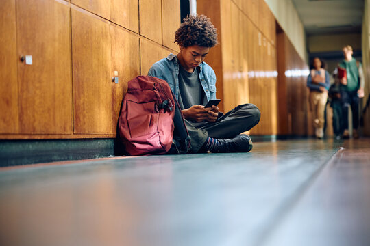 Black teenage boy texting on cell phone in hallway at high school.
