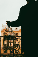 Silhouette of a young man in a coat holding a birdcage. Cityscape sunset on a background. - 718219433