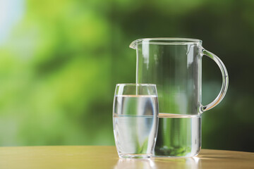 Jug and glass with clear water on table against blurred green background, closeup. Space for text