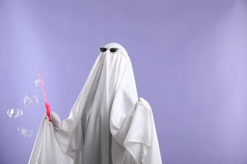 Funny ghost. Person in white sheet, sunglasses and soap bubbles on violet background, space for text