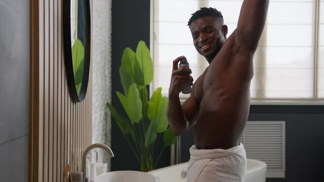 Happy African American man carefree dancing in home bathroom muscular sexy male guy looking in mirror applying deodorant in armpit fresh aroma antiperspirant sweat prevention body skin care hygiene