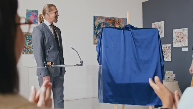 Medium shot of Caucasian male art gallery owner speaking to audience at exhibition opening, introducing young artist, who is removing cover and demonstrating new painting, and people clapping
