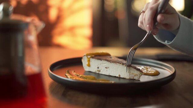 Woman eating a cheesecake slice sitting on the wooden table. Slice of tasty cake on plate pouring sauce