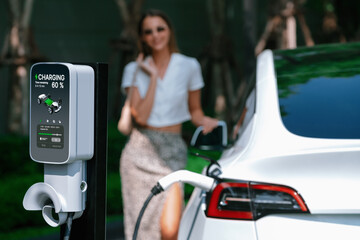Young woman travel with EV electric car charging in green sustainable city outdoor garden in summer. Urban sustainability lifestyle by green clean rechargeable energy of electric BEV vehicle innards - obrazy, fototapety, plakaty