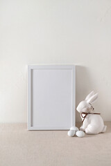 Empty white picture frame or poster mockup, white bunny decorative figurine, small eggs on neutral...
