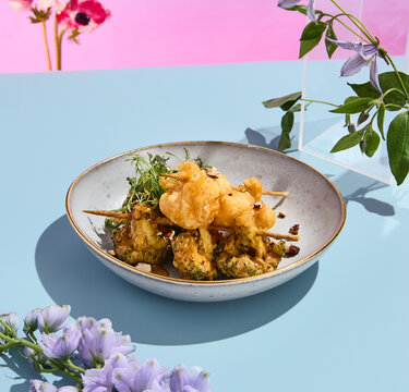 Elegant tempura vegetables on skewers with nut sauce amidst a surreal pastel floral backdrop