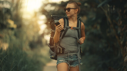 Young pretty white Caucasian girl with hiking backpack. Girl traveler smiles and holds a smartphone...