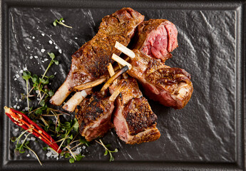 Juicy grilled lamb rack with a crusty sear, accented by fresh green herbs and a fiery red chili on a dark slate plate