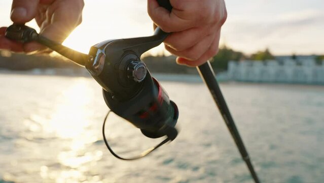 Man hobby fishing on sea tightens fishing line reel of fish summer. Handle rotation with reel of fishing rod against of orange sunset slow motion. Lens flare. Calm surface sea. Bright disk of sun