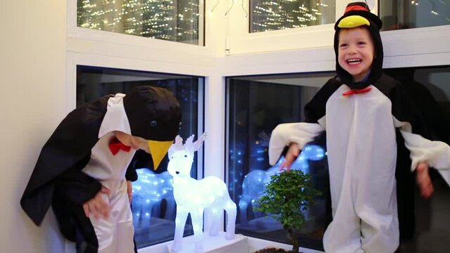 Two little boys in penguin costume are playing near deer and window.