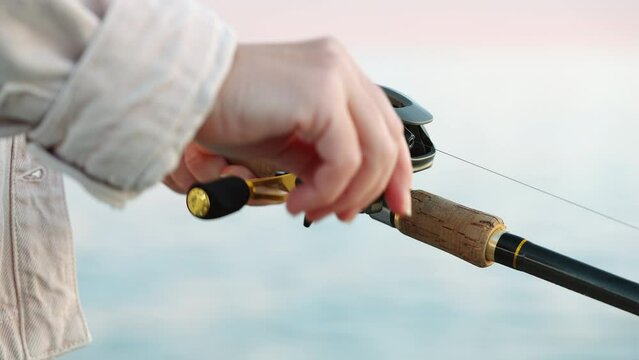 Reel of fishing rod hand rotation on blue cloudy sky of orange sunset slow motion. Woman hobby fishing on sea tightens fishing line reel fish summer. Go Everywhere. Lifestyle