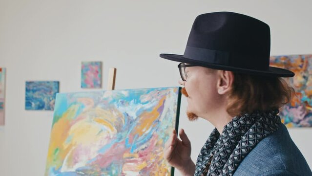 Close-up shot of eccentric young Caucasian man with red beard and moustache, in fedora hat, scarf, glasses describing abstract painting while giving public lecture on contemporary art styles in museum