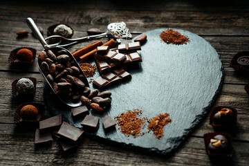 Handmade candies, two truffles, cocoa powder, chocolate and other lay on heart stone shape. Sweet delicious things on old wood background.