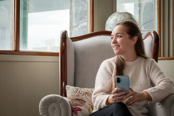 Mujer mirando por la ventana sonriente mientas usa su celular, sentada cómodamente en su sillón - obrazy, fototapety, plakaty